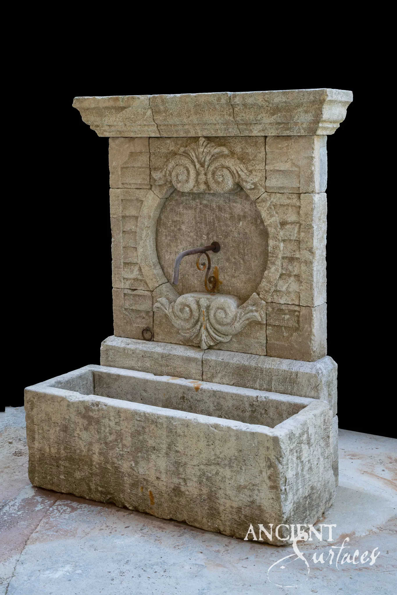 Reclaimed antique limestone wall fountain from Ancient Surfaces with a round medallion set in carved acanthus scrollwork over a rectangular trough basin