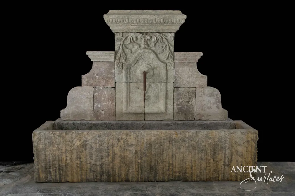 Reclaimed antique limestone wall fountain from Ancient Surfaces with a capital crowned backplate over a wide trough basin cut from banded grey and tan stone