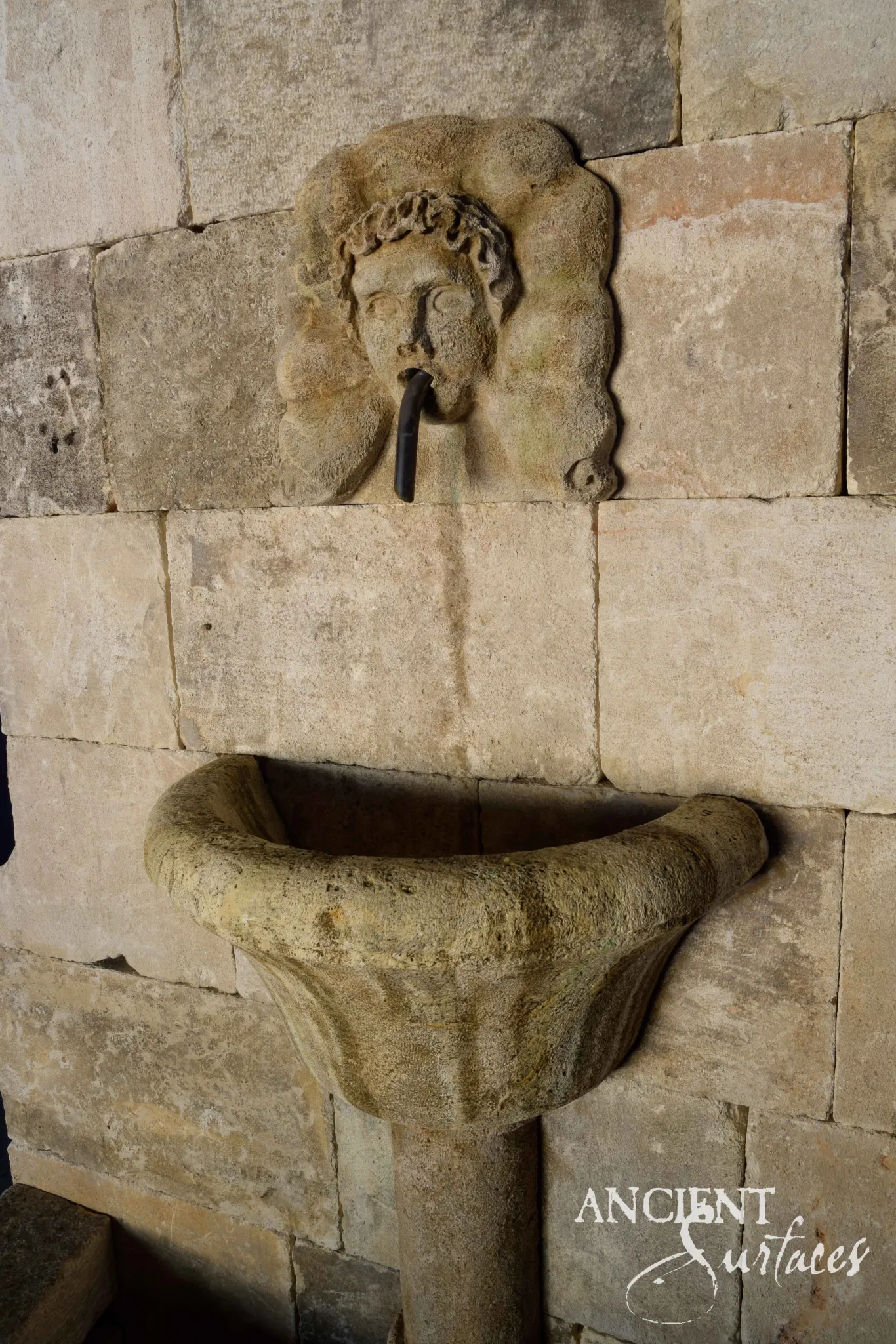 Detail of a reclaimed antique limestone lion mask wall fountain from Ancient Surfaces set flush into coursed ashlar stonework beneath a plain arched cresting