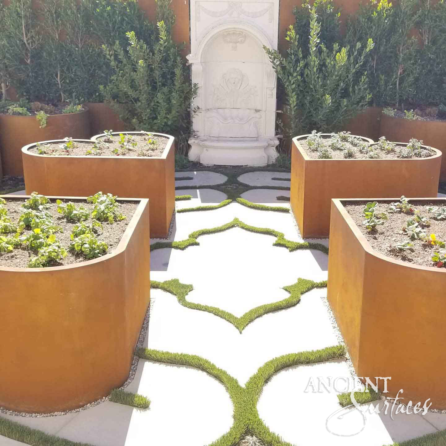 Reclaimed antique limestone wall fountain from Ancient Surfaces centred in a formal boxwood courtyard between raised corten steel planters and quatrefoil paving