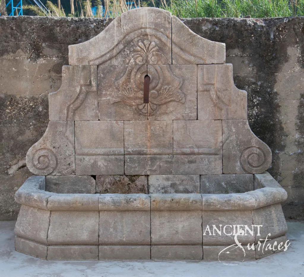 Reclaimed antique limestone wall fountain from Ancient Surfaces with a carved lion mask spout set into a rectangular niche in an ashlar garden wall beside a lawn