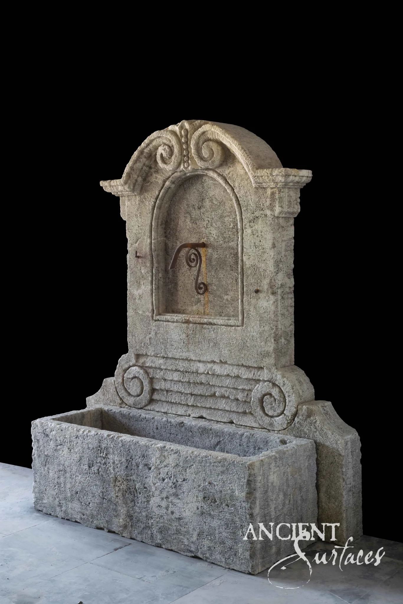 Reclaimed antique limestone wall fountain from Ancient Surfaces with a gadrooned scalloped vessel basin set between stone piers in a planted courtyard