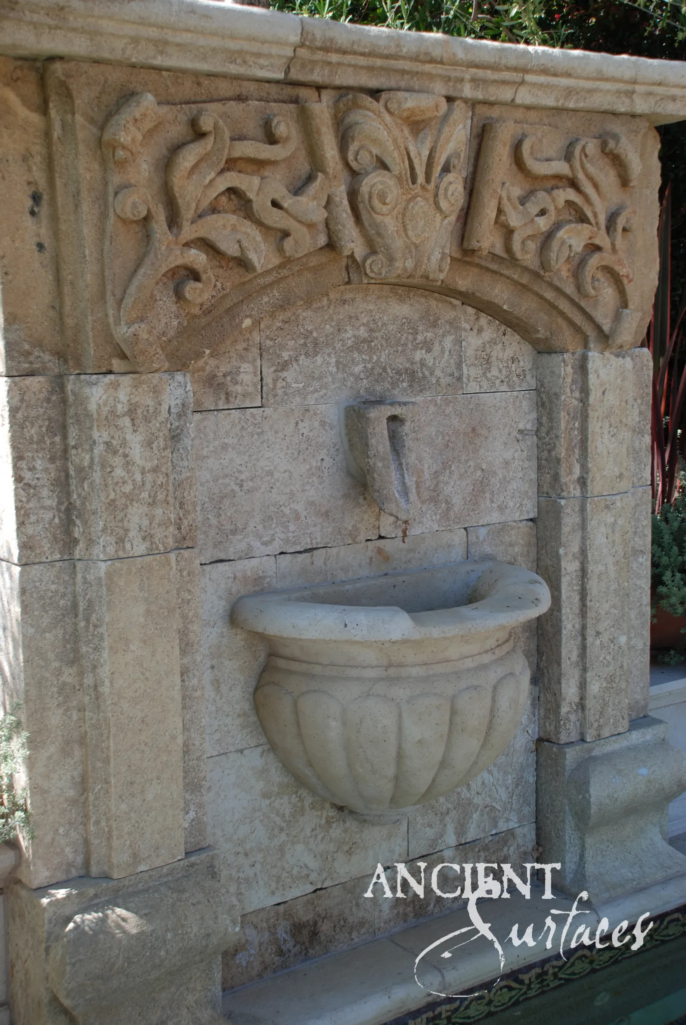 Reclaimed antique limestone wall fountain from Ancient Surfaces with a scrollwork crested pediment over a deeply gadrooned shell shaped basin bowl